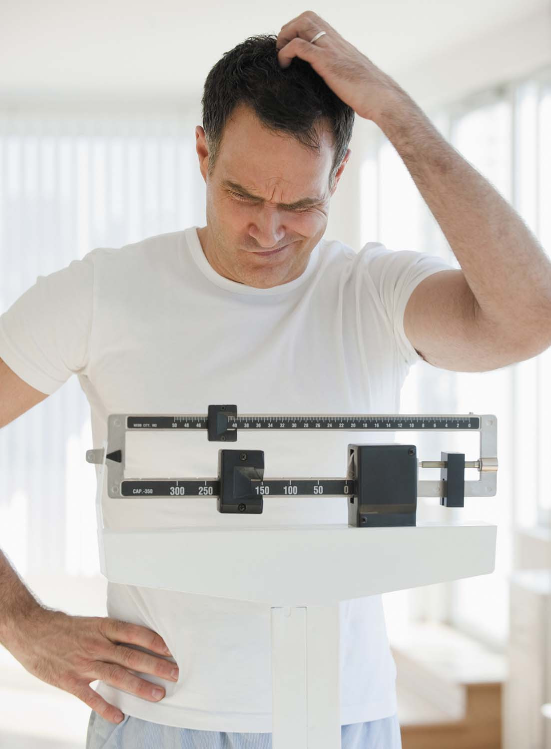 photo of a man standing on a balance-type scale and scratching his head as he looks at the number