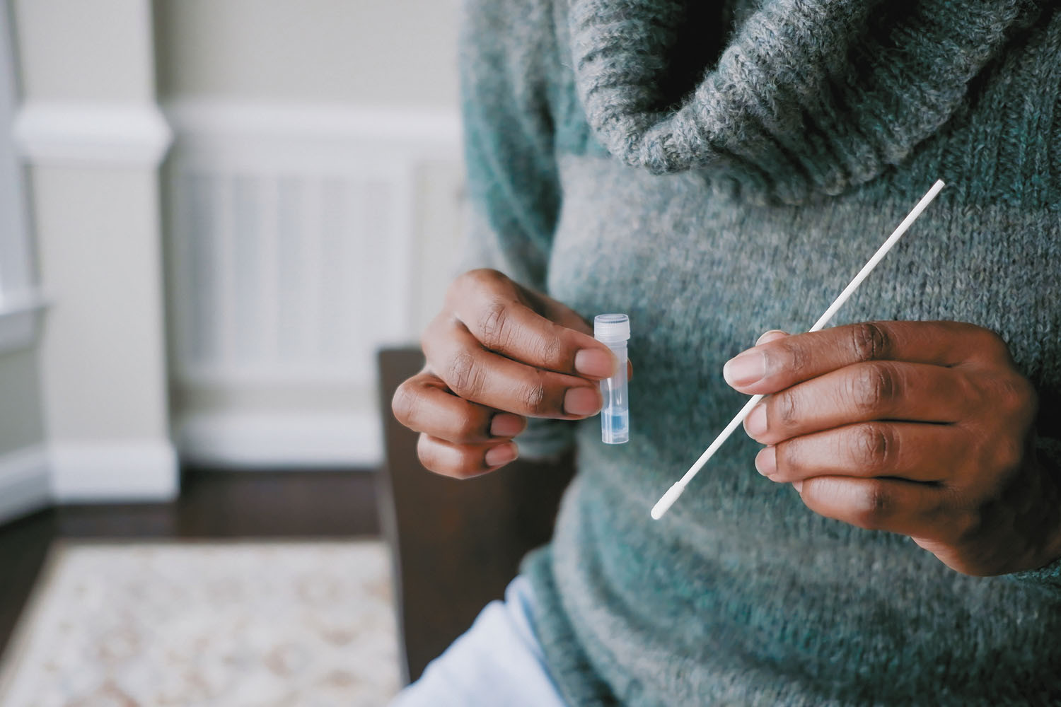 cropped photo of the torso and hands of a woman taking a home test for COVID-19; she is holding a nasal swab in one hand and the collection vial in the other