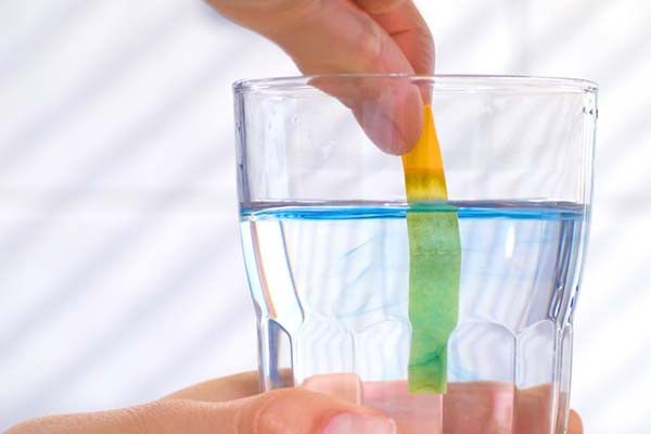 close-up photo of a hand holding a glass of water; the person's other hand is holding a paper strip in the water to test its pH level, and the part of the strip in the water is green
