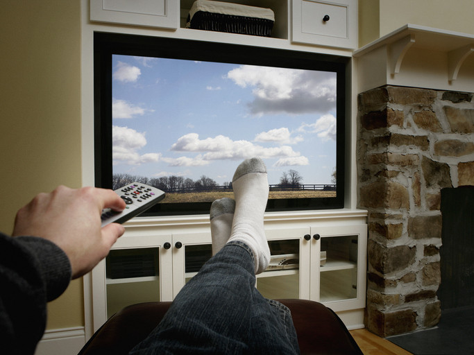 A close up of man's hand pointing a TV remote and sock-clad feet and legs in denim jeans up on a couch with TV in background showing beautiful blue skies, trees, and puffy clouds outside