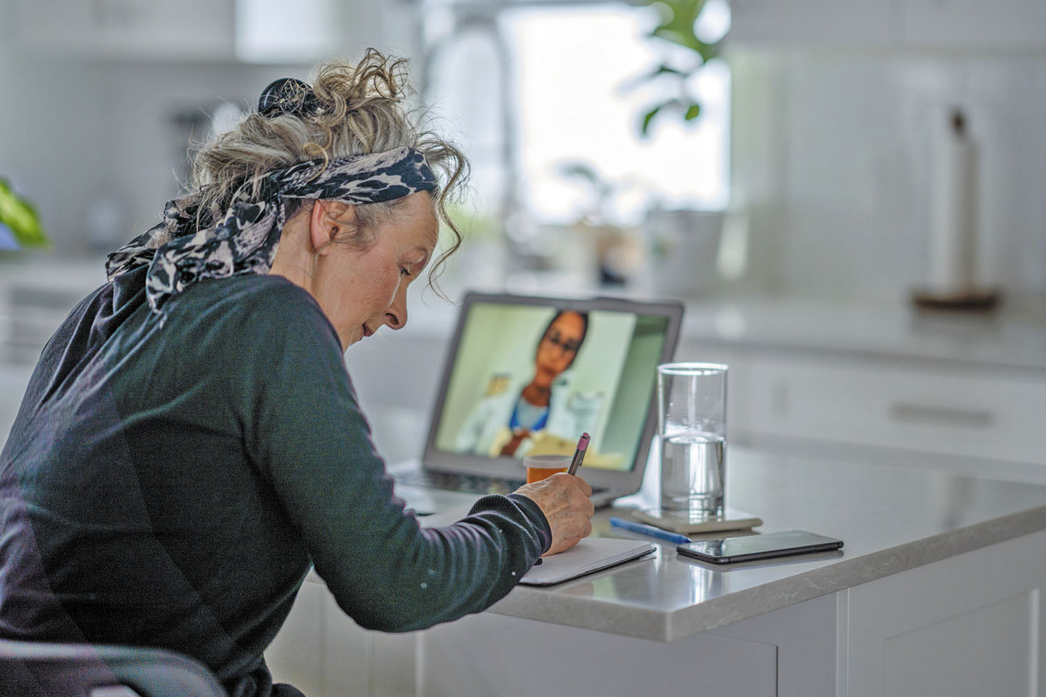 photo of a woman sitting at a table taking notes during a telehealth appointment; image of her doctor can be seen on a tablet propped up in front of her