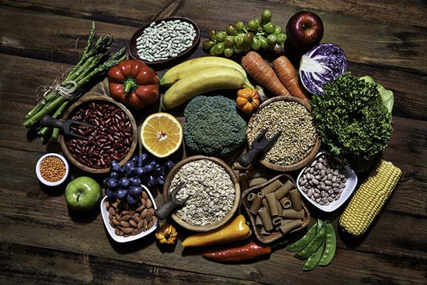 Overhead view photo of an assortment of fresh vegetables and legumes on a rustic wooden table, including corn, avocado, broccoli, orange, grapes, bell pepper, lettuce, banana, apple, almonds, and whole-grain pasta.