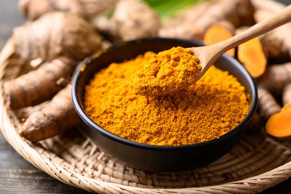 close-up photo of a bowl of turmeric powder with a wood spoon holding a generous amount; in the background out of focus is some fresh turmeric