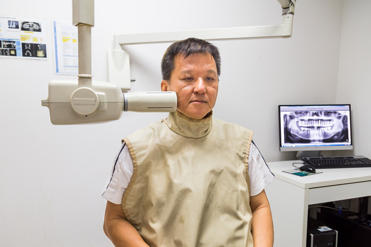 Man with black hair seated with x-ray machine pointed toward his jaw wearing lead protective vest; screen with xrays of teeth in backgroundvest 