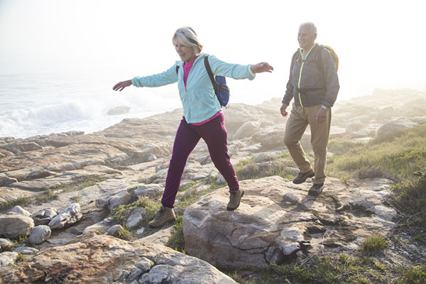 photo of a mature couple walking outdoors along a rocky coast; the woman has her arms extended out for balance as she steps from one rock to another