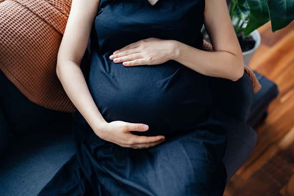 cropped photo showing the torso of a pregnant woman sitting on a couch holding her hands above and below her belly