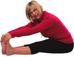 A mature woman sitting on the floor and stretching her arms out to onto her toes. 