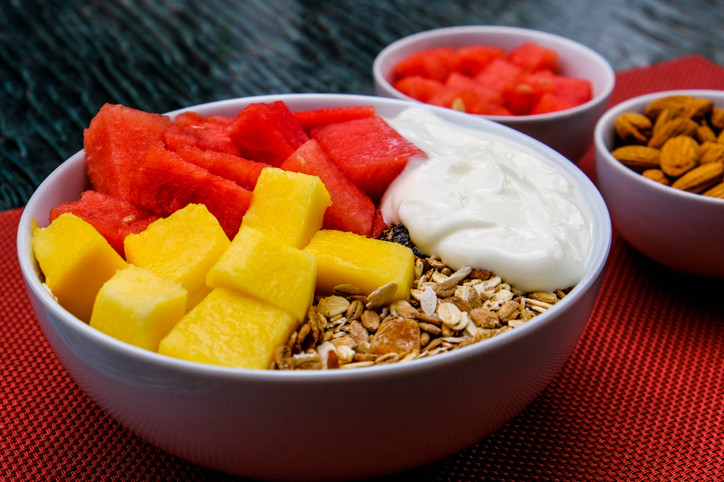 A bowl of whole-grain muesli, yogurt, red watermelon, and yellow mango with two little side bowls of nuts and fruit; concept is fiber and fermented foods