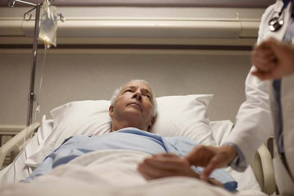 photo of a man in a hospital bed, he is looking to his left at a person who is partly visible but out of focus, with a hand on his wrist taking his pulse