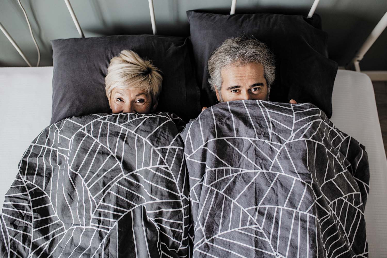 photo viewed from overhead of a couple in bed with the covers pulled up over the lower parts of their faces