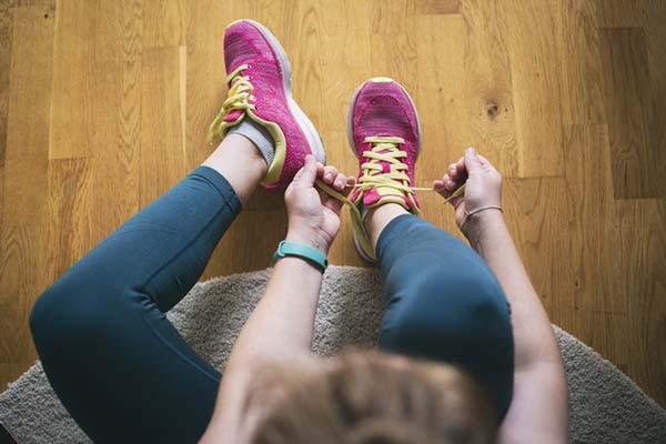 point-of-view photo looking down at the feet of a woman as she tightens the yellow laces on a raspberry-colored running shoe; also visible are a wood floor and her turquoise smartwatch strap