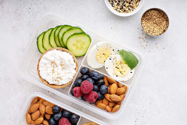 overhead view photo of a plastic lunch box containing high-protein foods including hard-boiled egg, berries, cucumber slices, cottage cheese, and nuts