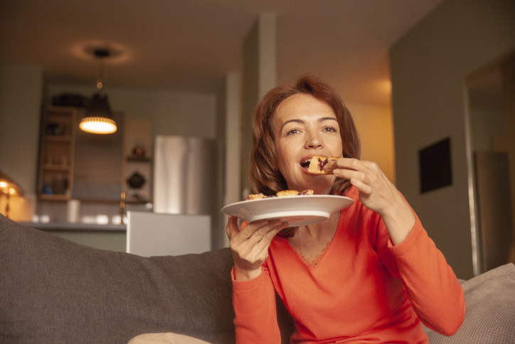 photo of a woman sitting in her couch holding a plate of cake with one hand and taking a bite from a piece she is holding with her other hand