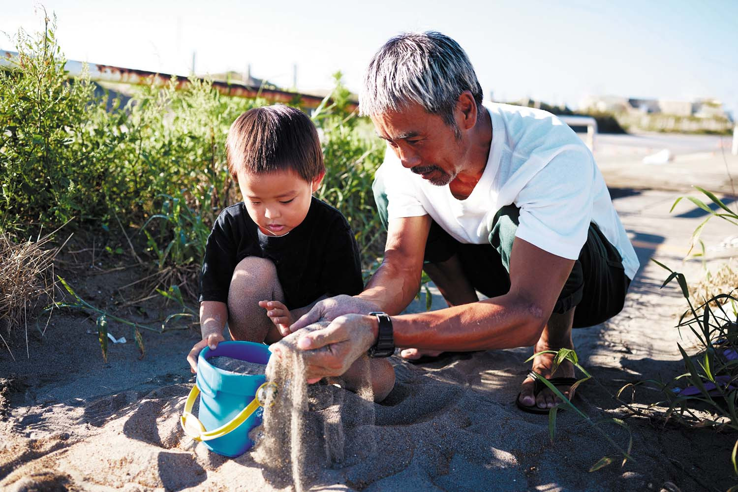 photo of a man doing a deep squat while playing with his grandchild; they have a bucket and are sifting sand on a beach