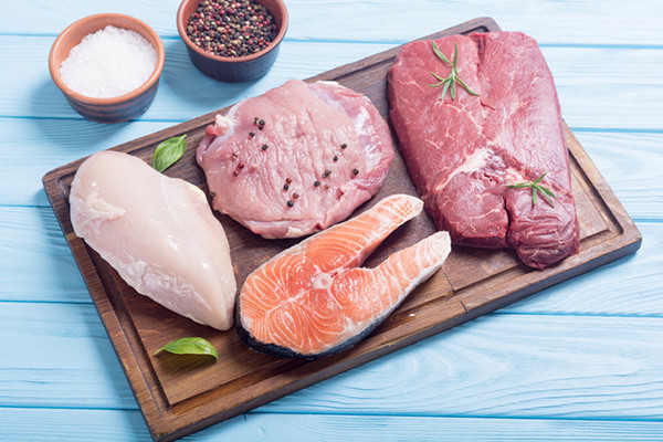 photo of a wood cutting board holding raw chicken, pork, salmon, and beef, with small bowls behind it holding salt and pepper