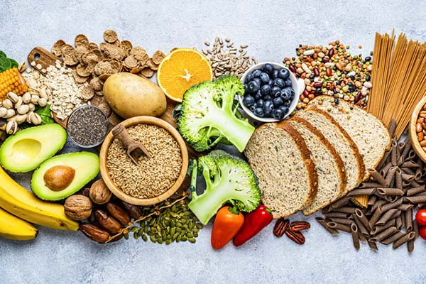 overhead view photo of an assortment of healthy foods, including bananas, corn avocado, orange, broccoli, blueberries, whole-grain bread, whole-wheat pasta, nuts, and seeds