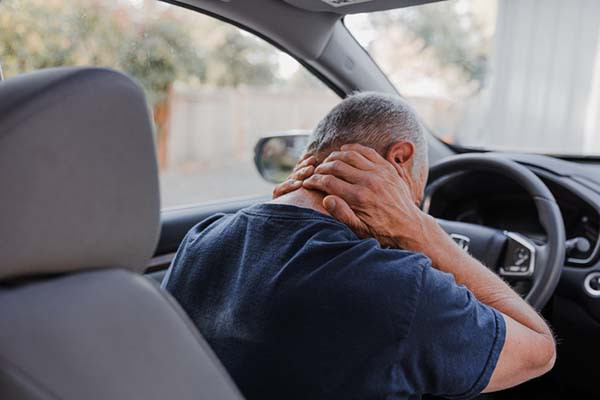 photo of a man in the driver's seat of a car, viewed from the back seat; he is hunched forward and is holding his hands on the back of his neck, indicating pain