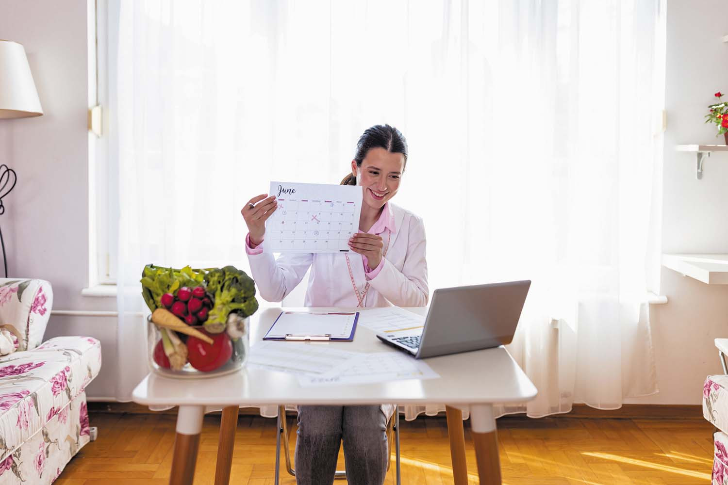 photo of a woman sitting at a small table, holding up a calendar page toward her laptop, suggesting she is in an oline meeting