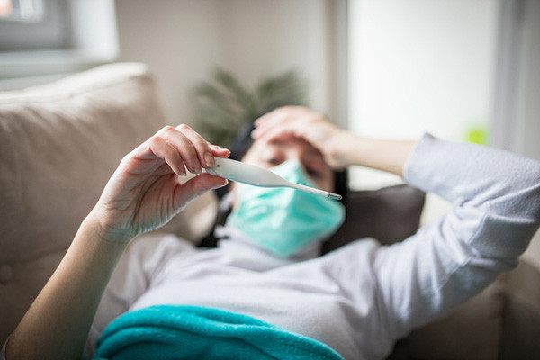 photo of a woman lying on a couch, holding up a thermometer to view the reading; her arm is in the foreground and her face is in the background out of focus, she is wearing a protective mask