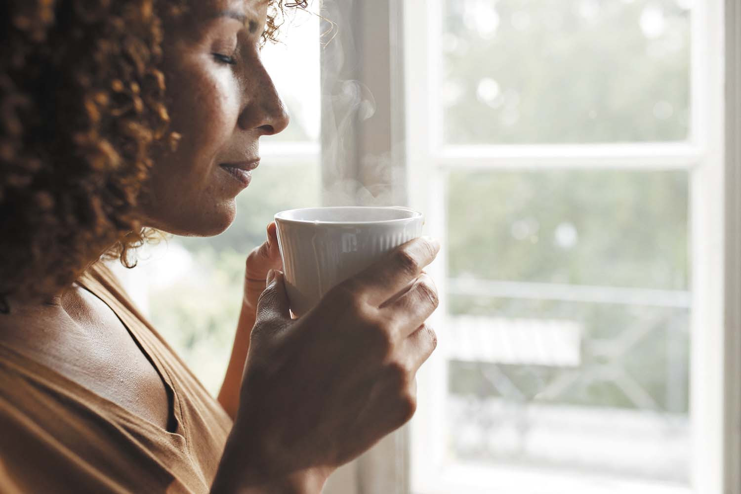 photo of a woman standing by a window while holding a cup of coffee