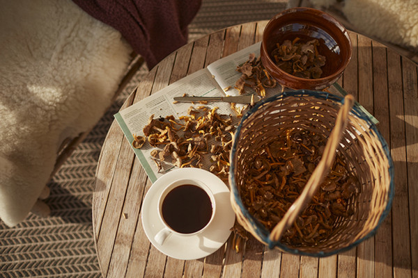 photo viewed from above of a cup of coffee on a saucer next to a basket and a bowl each holding freshly picked mushrooms; everything is on a small round wood table with additional loose mushrooms spread on an open magazine