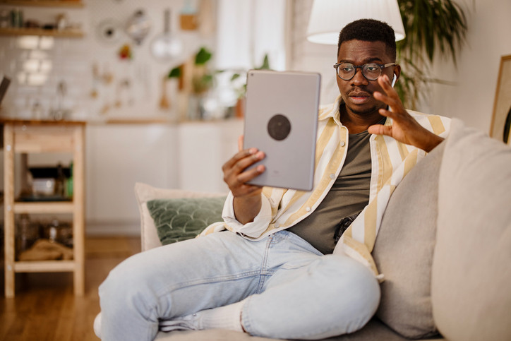 Young man dressed in yellow and white striped shirt on white couch holding tablet and gesturing during online mental health visit