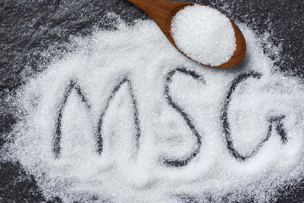 photo of a pile of monosodium glutamamte crystals on a dark textured surface with a small wooden scoop at the top; the letters M S G have been drawn into the pile by a finger