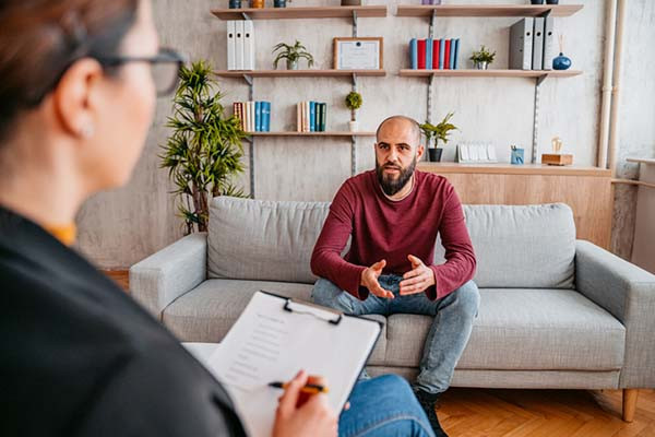 photo of a bearded man speaking while sitting on a sofa in a therapist's office; view is over the therapist's shoulder as she holds a clipboard with papers and a pen