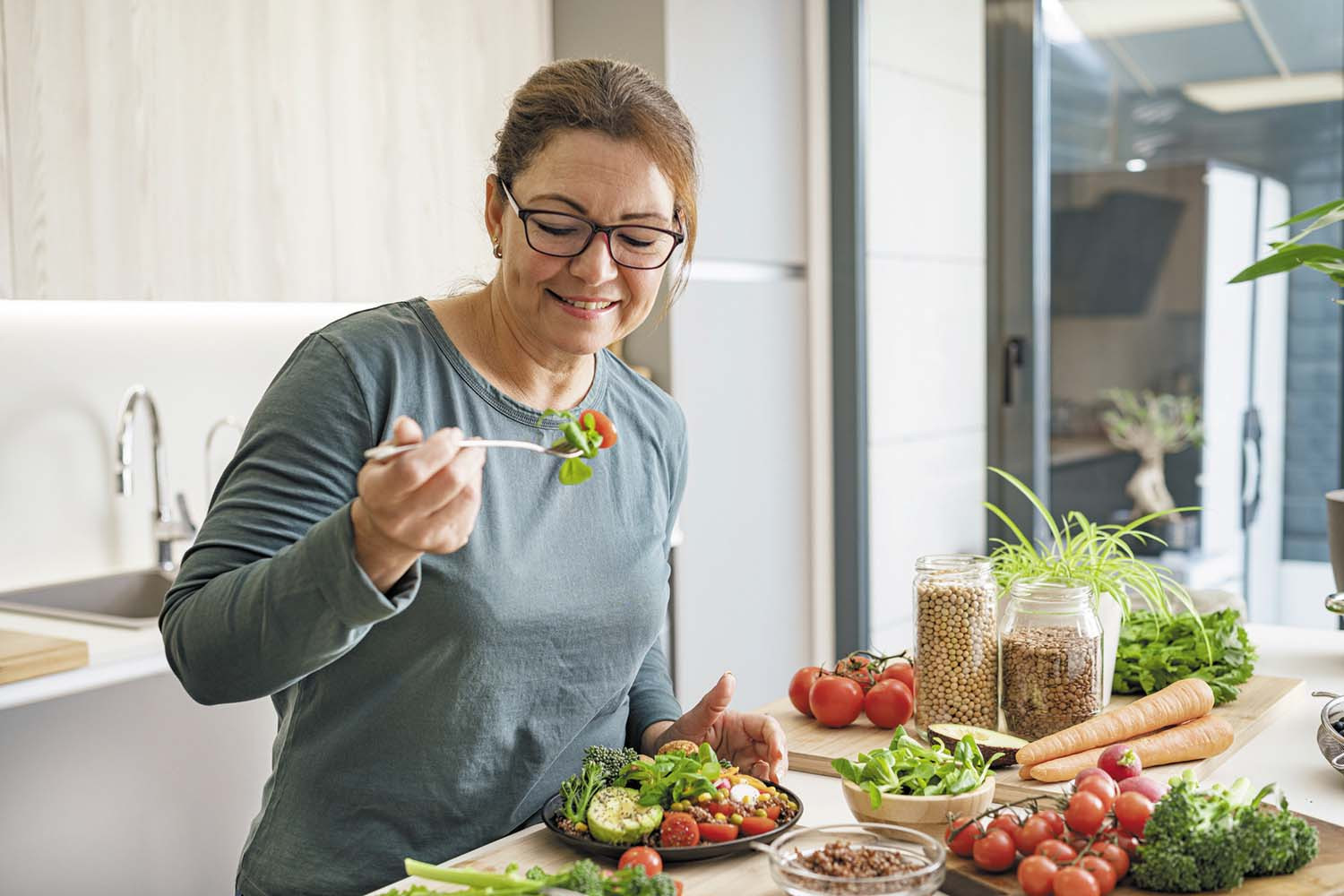 photo of a woman standing in front of a kitchen island that is filled with an assortment of healthy foods including fresh vegetables and jars of whole grains; she has prepared a salad and is holding a forkful, about to take a bite