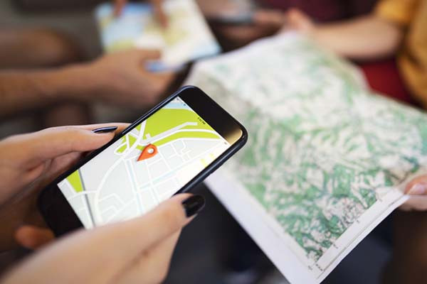 close-up photo of a woman's hands holding a smartphone that is displaying a map with a location marker; there is a paper terrain map open on the table under the phone