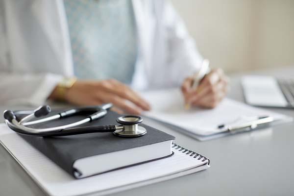 cropped photo showing a desk with two notebooks and a stethoscope resting on top; the torso of a figure is sitting at the desk, out of focus, wearing a white lab coat and writing notes