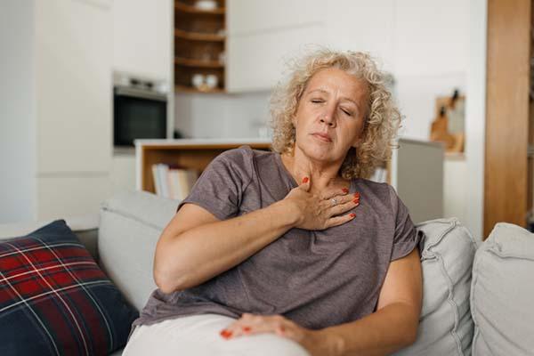 photo of a middle-age woman sitting on a sofa and experiencing chest pain, she has her eyes closed and her hand over her heart