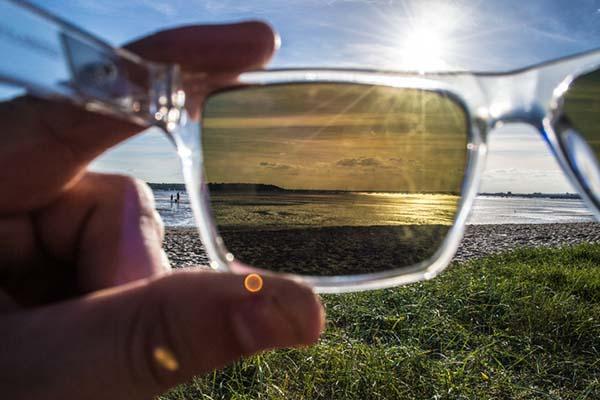 point of view photo of a hand holding a pair of sunglasses; the glasses have clear frames and brown lenses, and behind them is visible a grassy area then beach and ocean