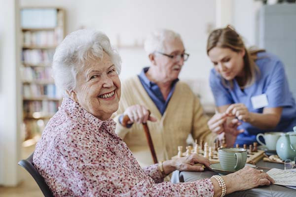 photo of a smiling senior woman sitting at a table with a cup and saucer in front of her; in the background, out of focus, is a health care worker assisting a senior man with a chess board in front of him