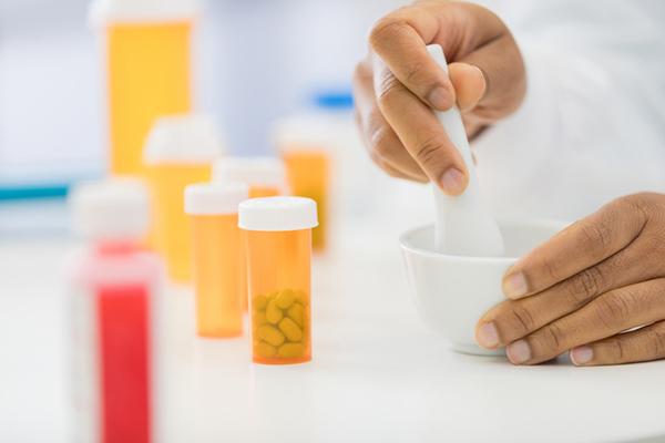 Cropped image showing the hands of a pharmacist using a mortar and pestle to prepare a medication, with several orange plastic bottles on the counter nearby, some empty and some containing pills.