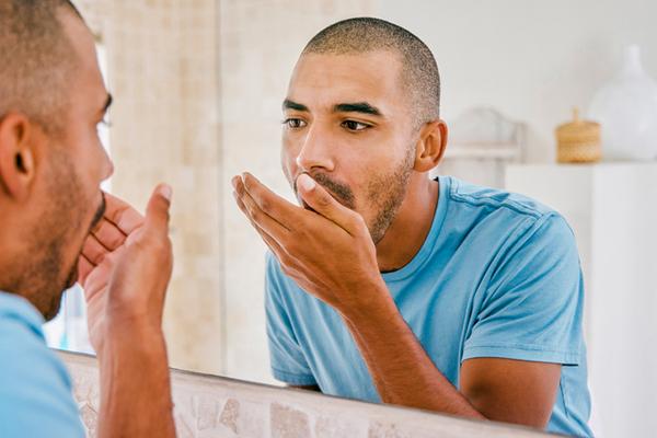 photo of a young man standing in front of a bathroom mirror and holding his hand in front of his mouth to check his breath