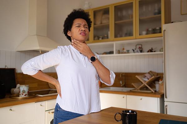 Woman standing in her kitchen, holding a hand to her throat as she experiences sore throat pain.