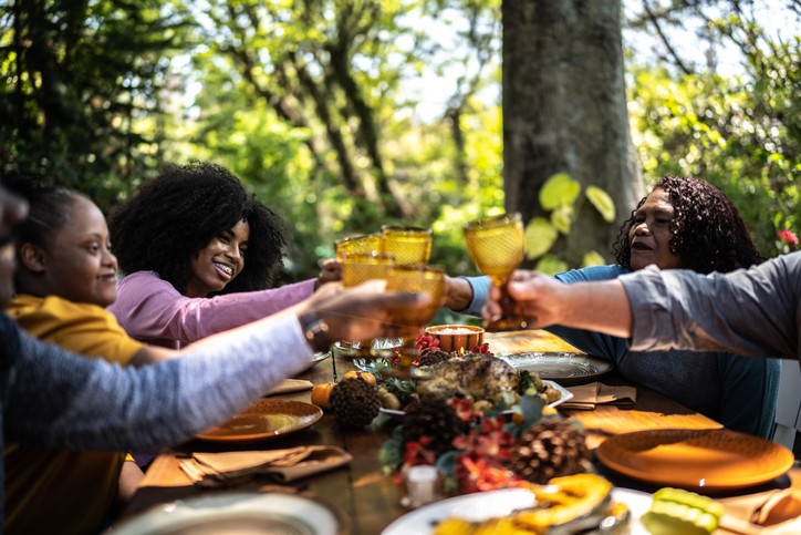 A family eating a meal near leafy green trees outdoors; three out of five people are seen close up during a toast
