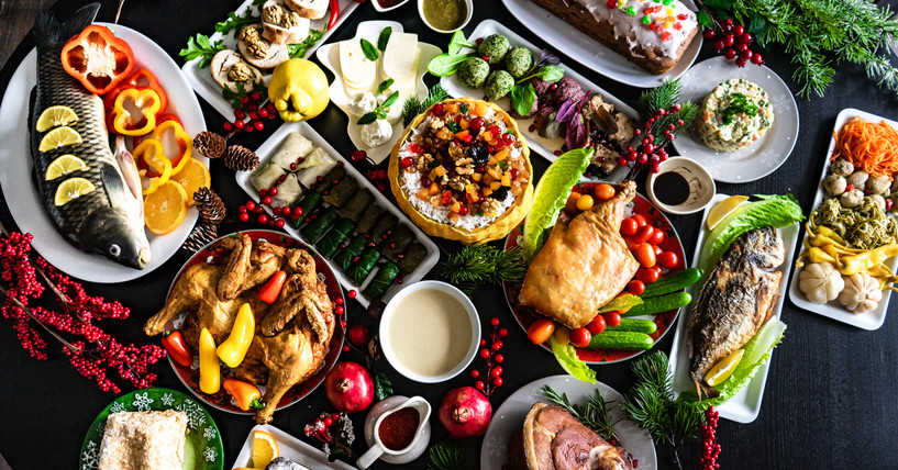 A table tightly packed with holidays foods: whole fish with slices of lemon, golden brown poultry, ham, colorful vegetables, cakes, blintzes, and more